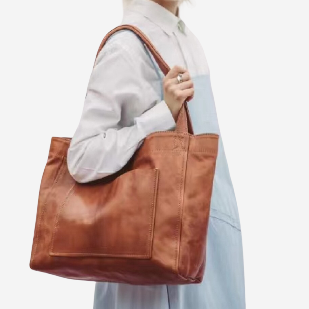 A person wearing a white shirt and blue dress is holding a brown leather tote bag.