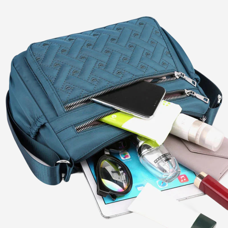 A blue crossbody bag with a quilted pattern, containing various personal items such as a smartphone, sunglasses, and a bottle of lotion.