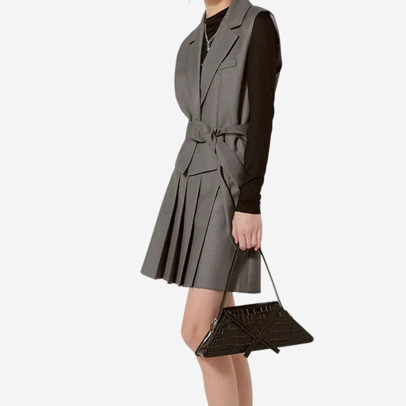 A person wearing a gray dress and carrying a black purse is standing against a white background.